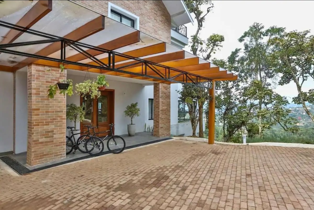 An outdoor area features a covered entrance with a wooden awning supported by metal beams. Two bicycles are parked beside the entrance, and potted plants add greenery to the space. The surrounding trees contribute to a natural setting.