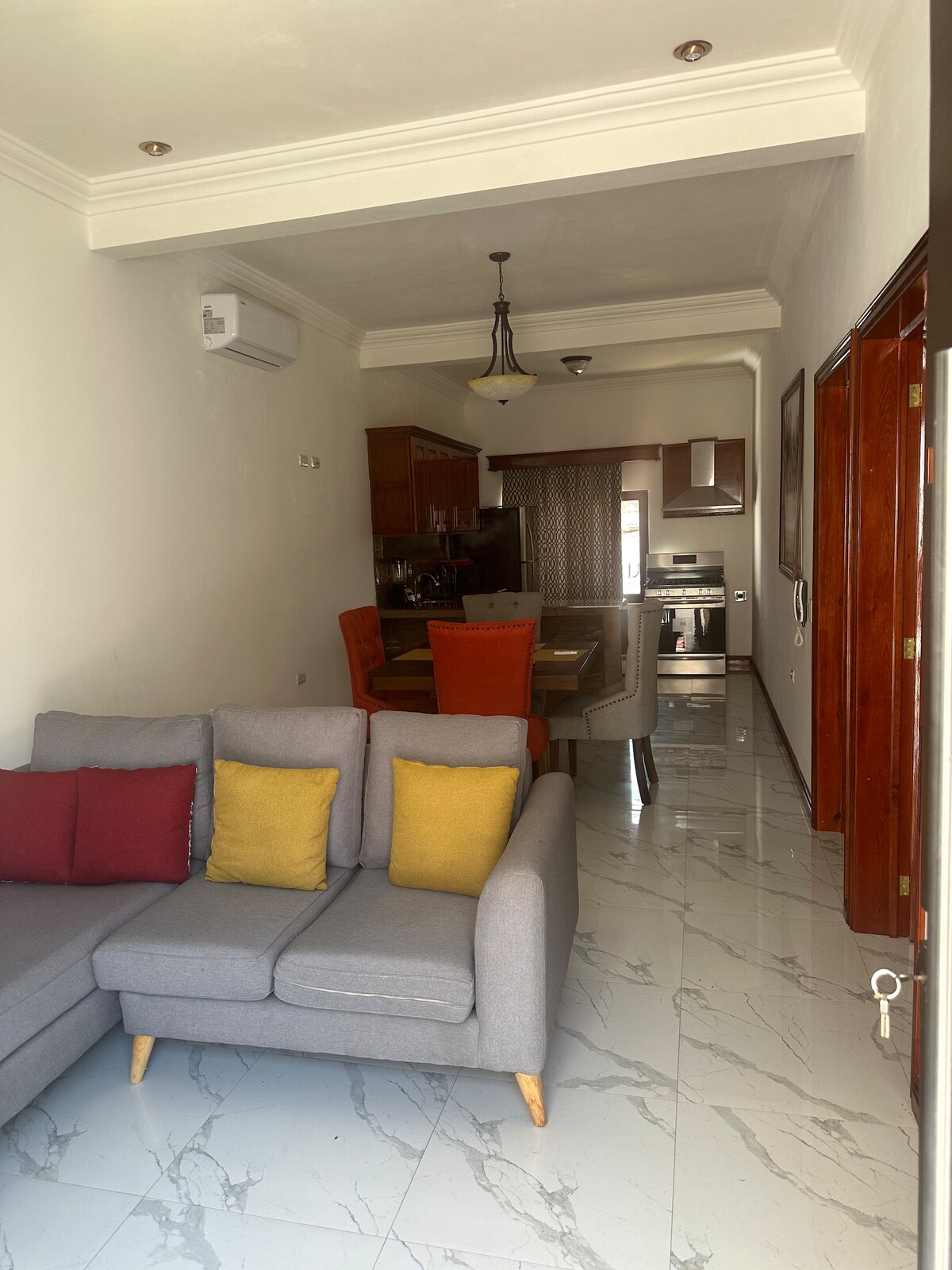 A bright living area features a gray sofa adorned with colorful cushions. A dining table with orange and gray chairs is visible in the background, alongside a modern kitchen equipped with stainless steel appliances. Glossy marble flooring complements the serene ambiance.
