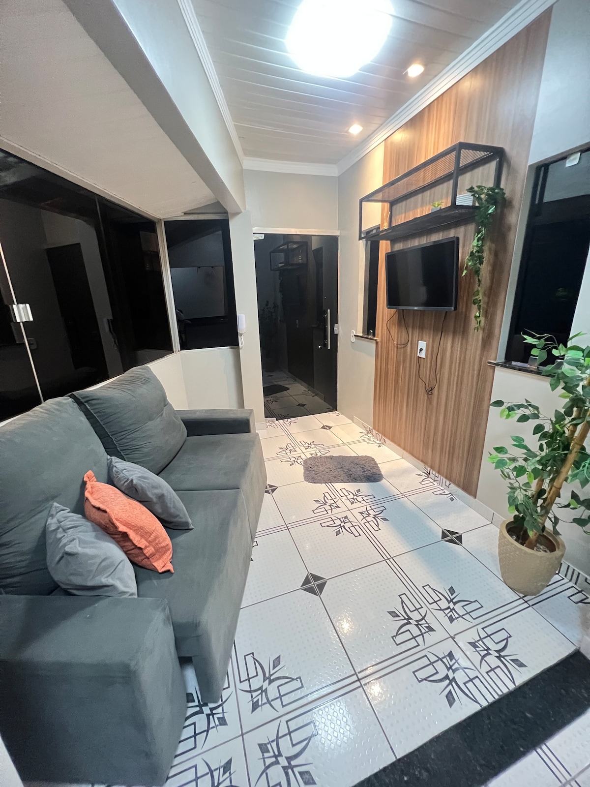 A compact seating area is presented with a gray couch adorned with decorative pillows. The walls feature a blend of light and wood accents, while a television is mounted above a slim console. Natural light filters through the windows, illuminating the patterned floor tiles.