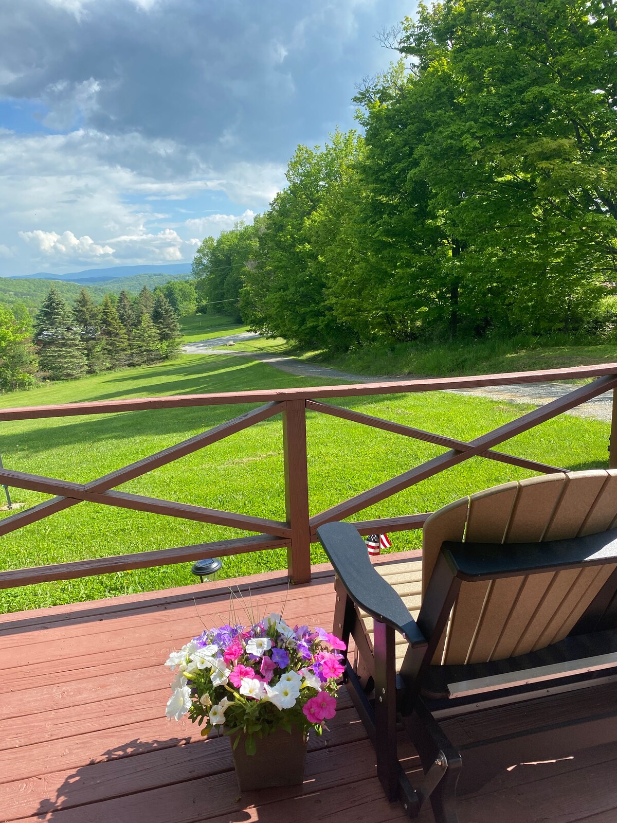A welcoming porch is featured, showcasing a comfortable chair and a planter filled with colorful flowers. Lush greenery and trees are visible in the distance, complementing the scenic view of the Endless Mountains under a partly cloudy sky.