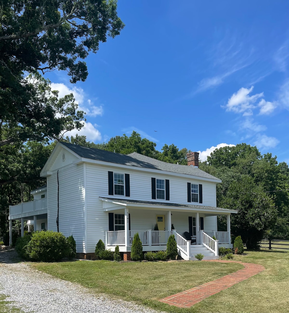 A charming two-story farmhouse is depicted, featuring a white exterior with black shutters. A wide porch extends across the front, adorned with white railings. A brick pathway leads through the manicured lawn, surrounded by greenery under a bright blue sky dotted with clouds.