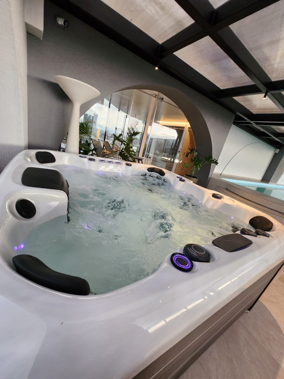 A modern jacuzzi is featured with bubbles gently rising, illuminated by soft LED lights. Surrounding plants enhance the calm ambiance, while a large mirror reflects the serene space. The jacuzzi is positioned near expansive windows, offering a view of the outside environment.