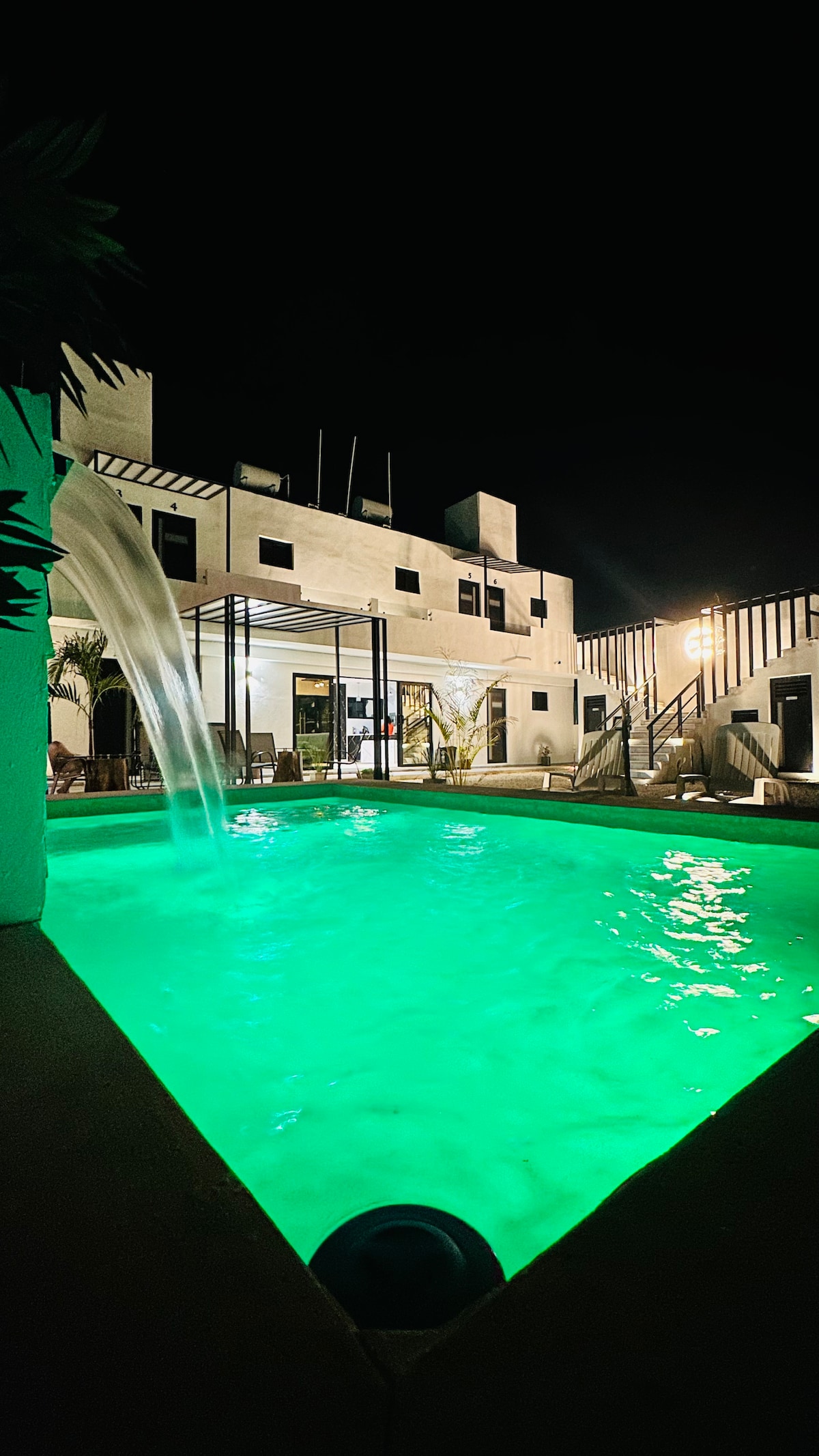 The pool area is illuminated in a soft green hue, complemented by a cascading waterfall. The house features a modern architectural design with large windows reflecting the ambient lighting, offering a glimpse of the inviting outdoor space.