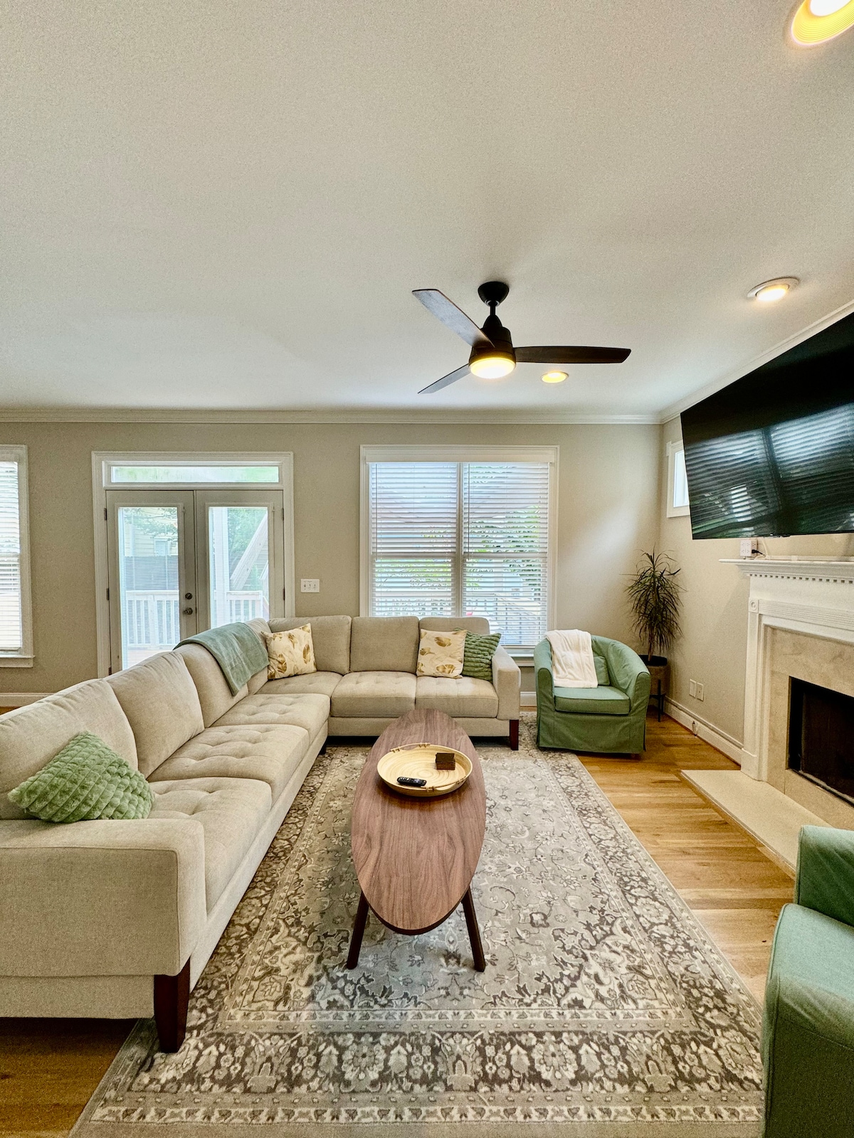 A comfortable living area features a neutral sectional sofa and two armchairs. A wooden coffee table with decorative items sits centrally on a patterned area rug. Natural light filters through the windows, illuminating the space that includes a television mounted above the fireplace.