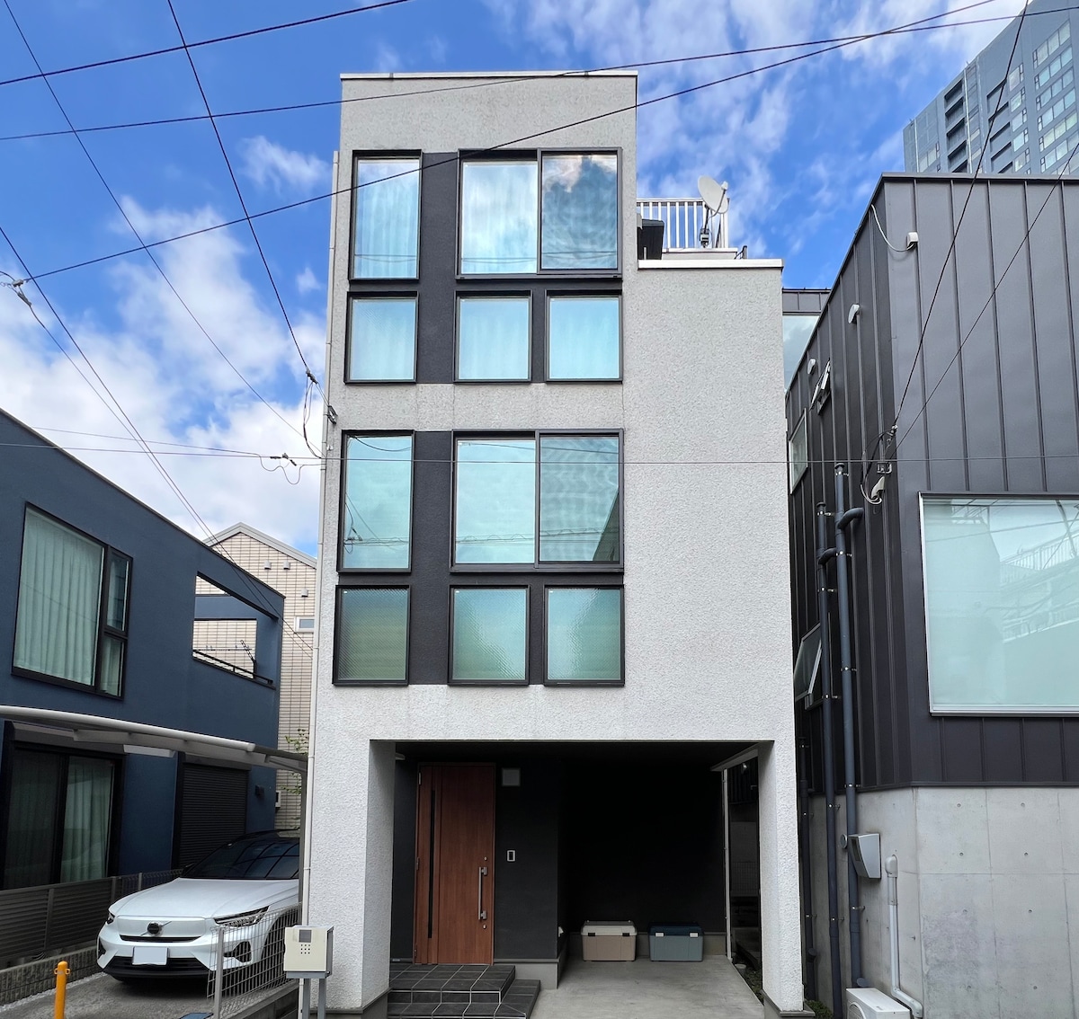 The exterior of a three-story house is presented, featuring a modern design with multiple large windows in the upper section. A compact parking area is visible in front, with a sleek entryway leading to the main door. The sky above is clear with clouds.