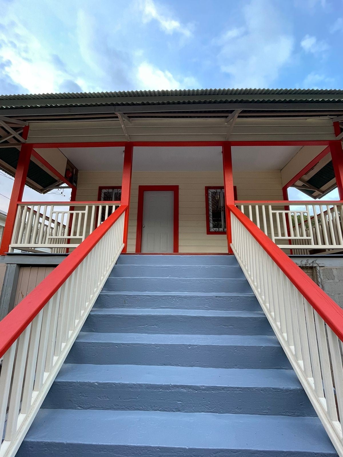 A staircase with white railings leads to a welcoming entrance, framed by red accents. The steps are painted in light blue, and a simple white door is positioned at the top, inviting guests inside.