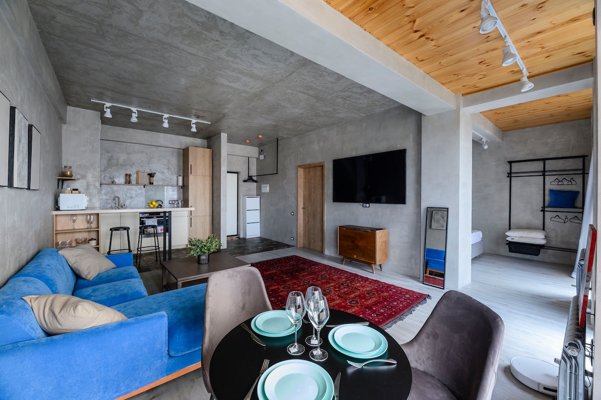 An open living space features a blue sofa and a round dining table set for four. A mounted television is positioned on a concrete wall, while a kitchenette is visible in the background. Soft lighting and wooden beams above add warmth to the modern design.