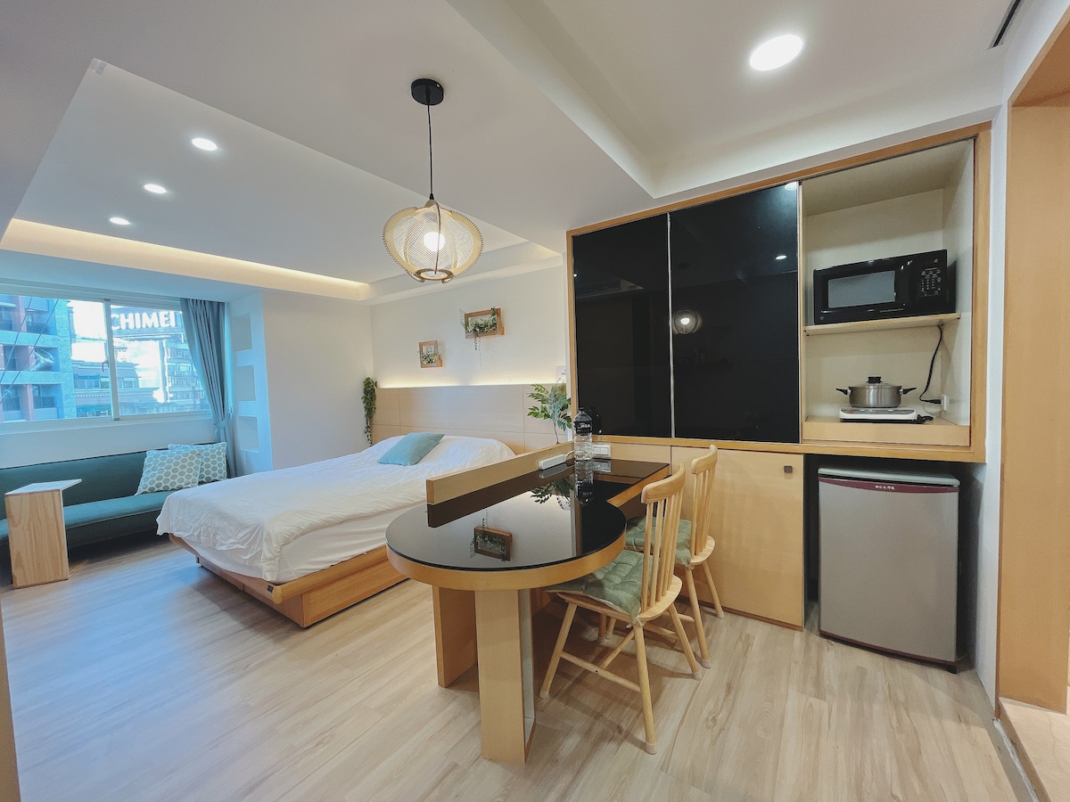 A spacious studio apartment features a comfortable bed and a dining table with chairs. A kitchenette is equipped with a microwave and cooking utensils. Natural light fills the room through a large window, while a comfortable seating area is visible in the background.