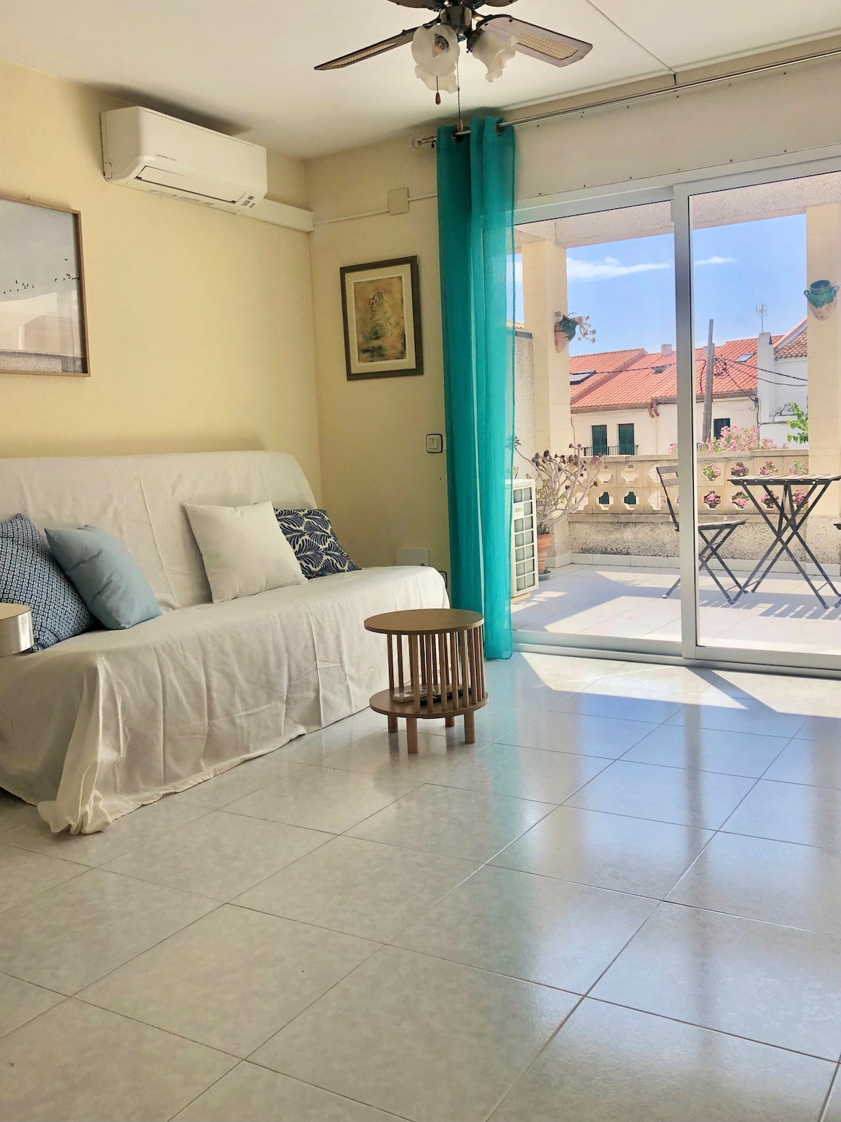 A light-filled living area is presented, featuring a sofa covered with a white throw, alongside a small wooden table. A fan hangs from the ceiling. Large glass doors open to a balcony with views of nearby rooftops, and a light blue curtain gently frames the doorway.