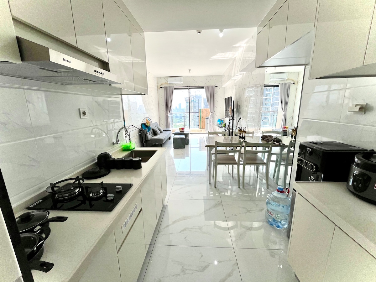 A modern kitchen is shown, featuring sleek cabinetry and a gas stove. Natural light floods the space, highlighting a dining area with a table and chairs. In the background, a spacious living room can be seen, with a large window offering a view of the cityscape.