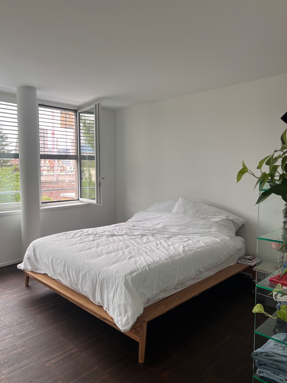 A bright bedroom features a spacious bed with white bedding against a light wall. Large windows provide natural light and a view of greenery outside. A modern shelf with plants and personal items is located beside the bed, enhancing the room's fresh ambiance.