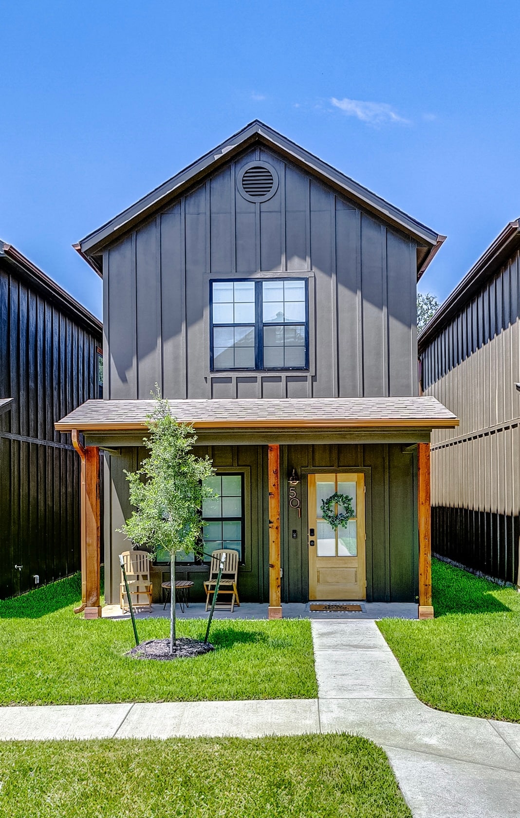 A modern cottage home is featured with a dark exterior, showcasing a welcoming front porch. Two chairs sit beside a small tree, enhancing the outdoor space. A well-maintained lawn and a concrete pathway lead to the door, complemented by a single window above the entrance.