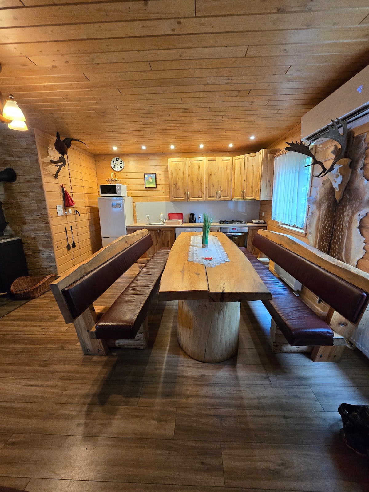 A spacious dining area features a large wooden table surrounded by long benches. The warm wooden walls add to the inviting feel, while overhead lighting creates a bright environment. A modern kitchen is visible in the background, enhancing the functionality of the space.
