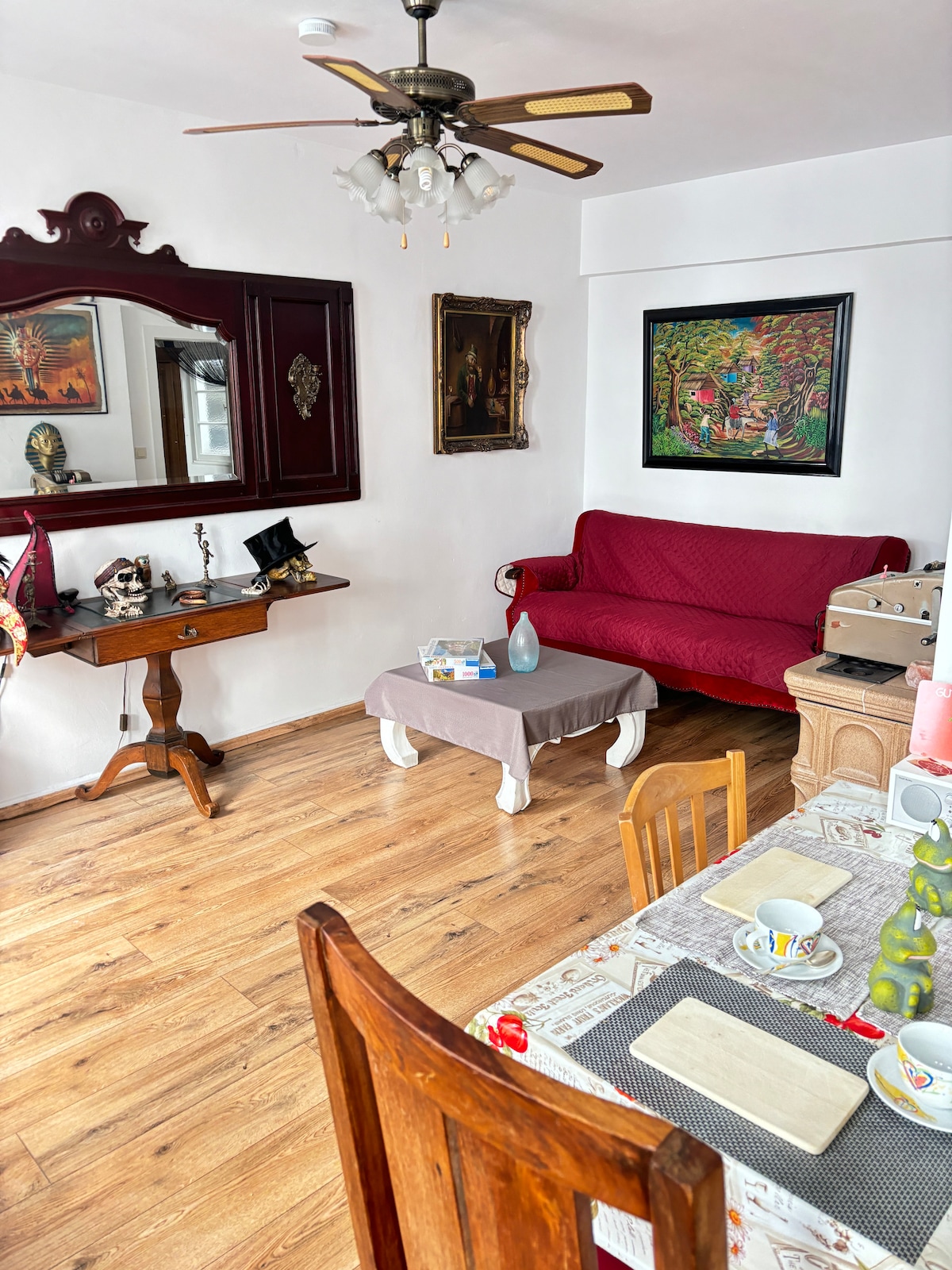 A living area features a red sofa, a wooden table with chairs, and an elegant console table. Artwork adorns the walls, providing a touch of color. The space is well-lit with natural light, accentuated by a ceiling fan and wooden flooring.