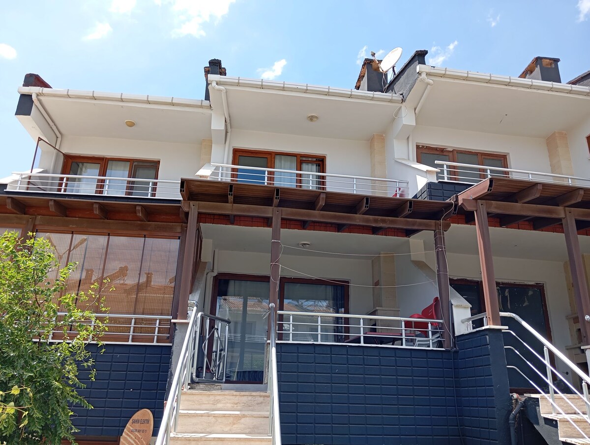 A three-story villa is presented, featuring balconies with glass railings. A staircase leads up to the main entrance, flanked by greenery. The exterior includes a mix of stone and wood, highlighting the modern design under a clear blue sky.