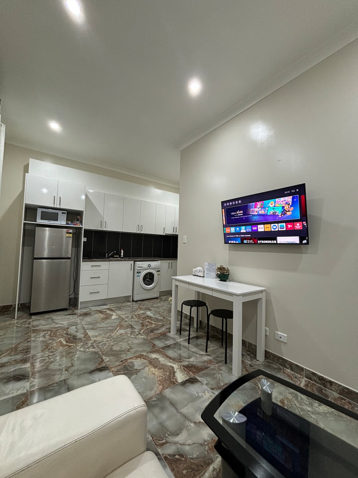 A modern kitchen area is visible, featuring sleek white cabinets and stainless steel appliances, including a refrigerator and washing machine. A small dining table with two chairs is positioned near the wall-mounted TV, creating a functional and open living space.