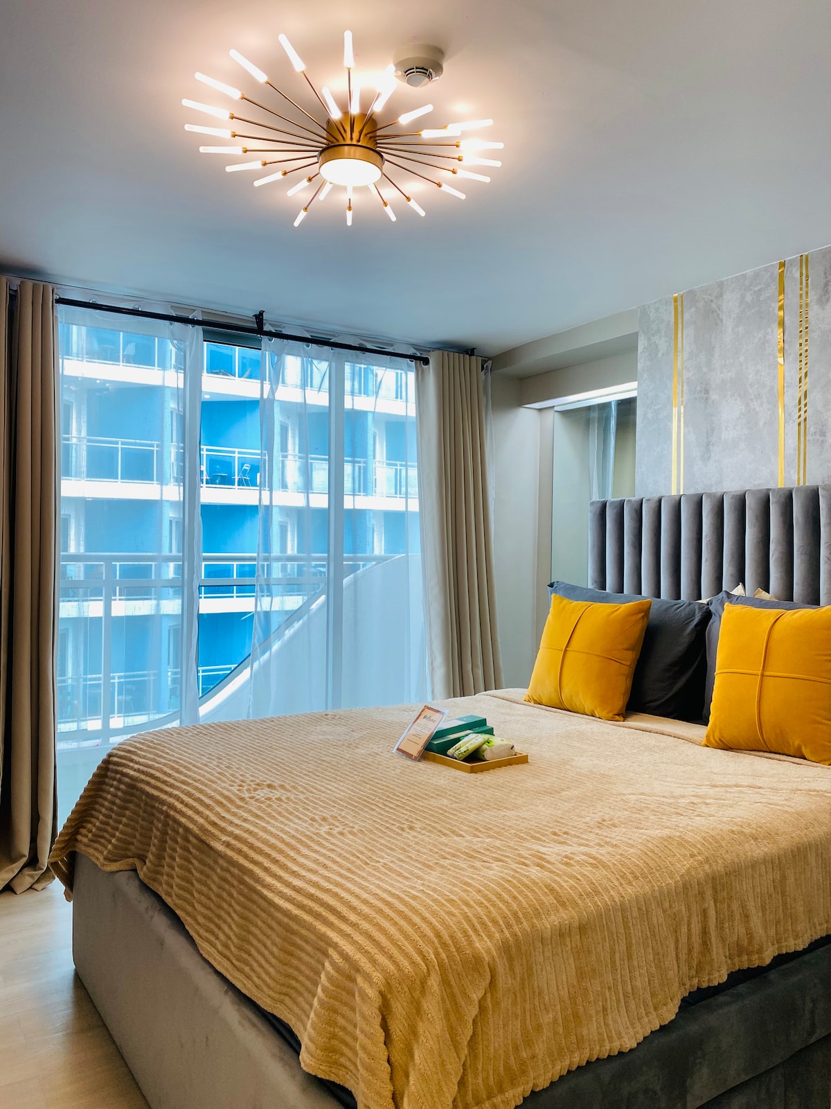 A cozy bedroom features a spacious bed adorned with muted striped linens and two decorative pillows. Natural light enters through large, sheer curtains covering a glass door, which opens to a balcony. A modern light fixture hangs above, complementing the serene atmosphere.