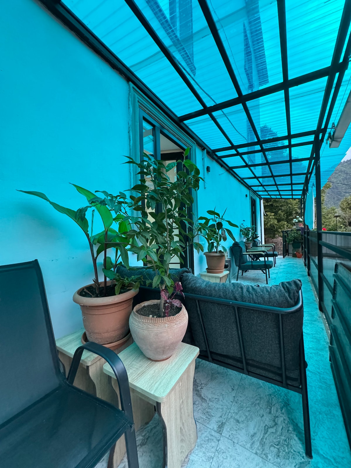 A spacious terrace is shown, featuring potted plants and comfortable seating arrangements. A table is placed beside the seating area, while a canopy of blue-tinted glass provides shade. The terrace extends alongside the building, inviting relaxation in a serene setting.