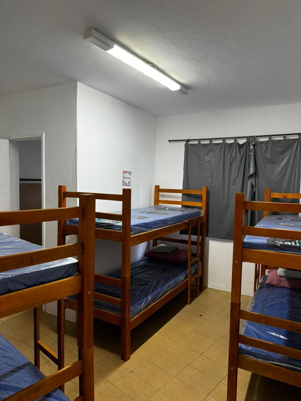 A shared room is equipped with two sets of sturdy wooden bunk beds, each with a blue mattress and a pillow. A large window is covered by dark grey curtains, and a light fixture hangs from the ceiling, illuminating the space.