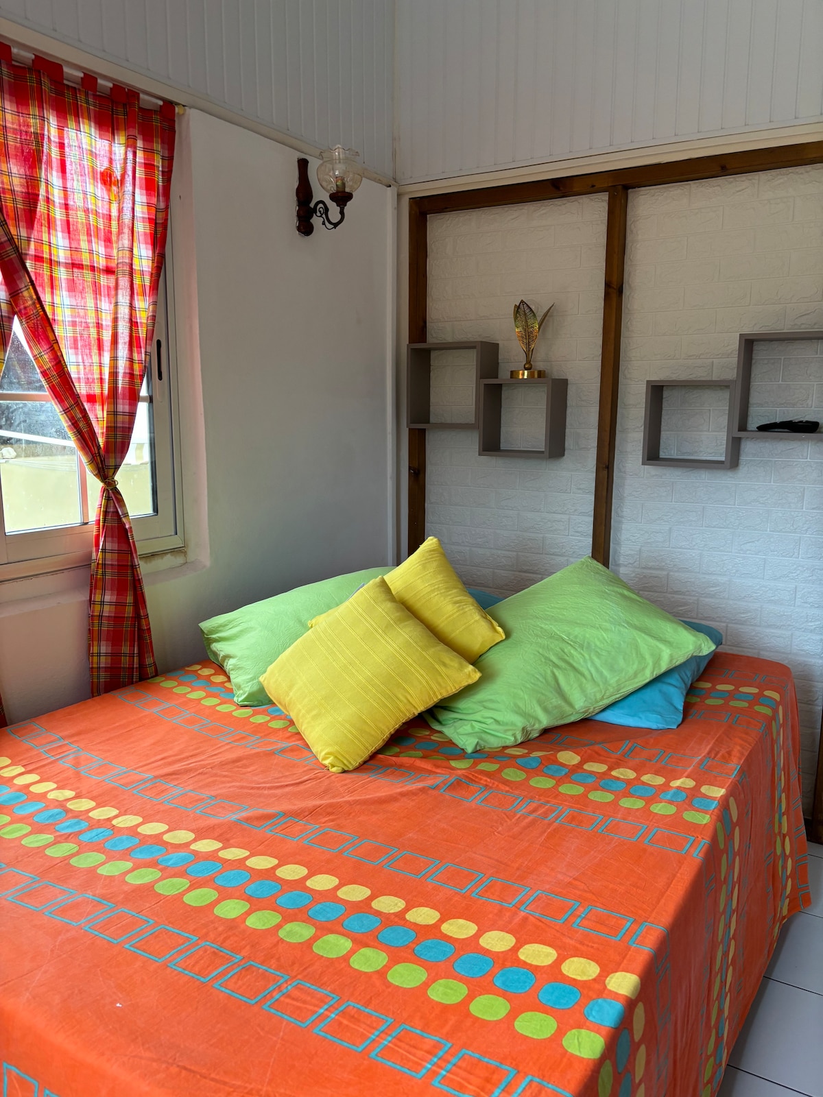 A colorful sleeping area features a bed adorned with an orange patterned coverlet. Several cushions in green and yellow are arranged casually. Bright red and white checked curtains frame the window, adding a cheerful touch to the space.