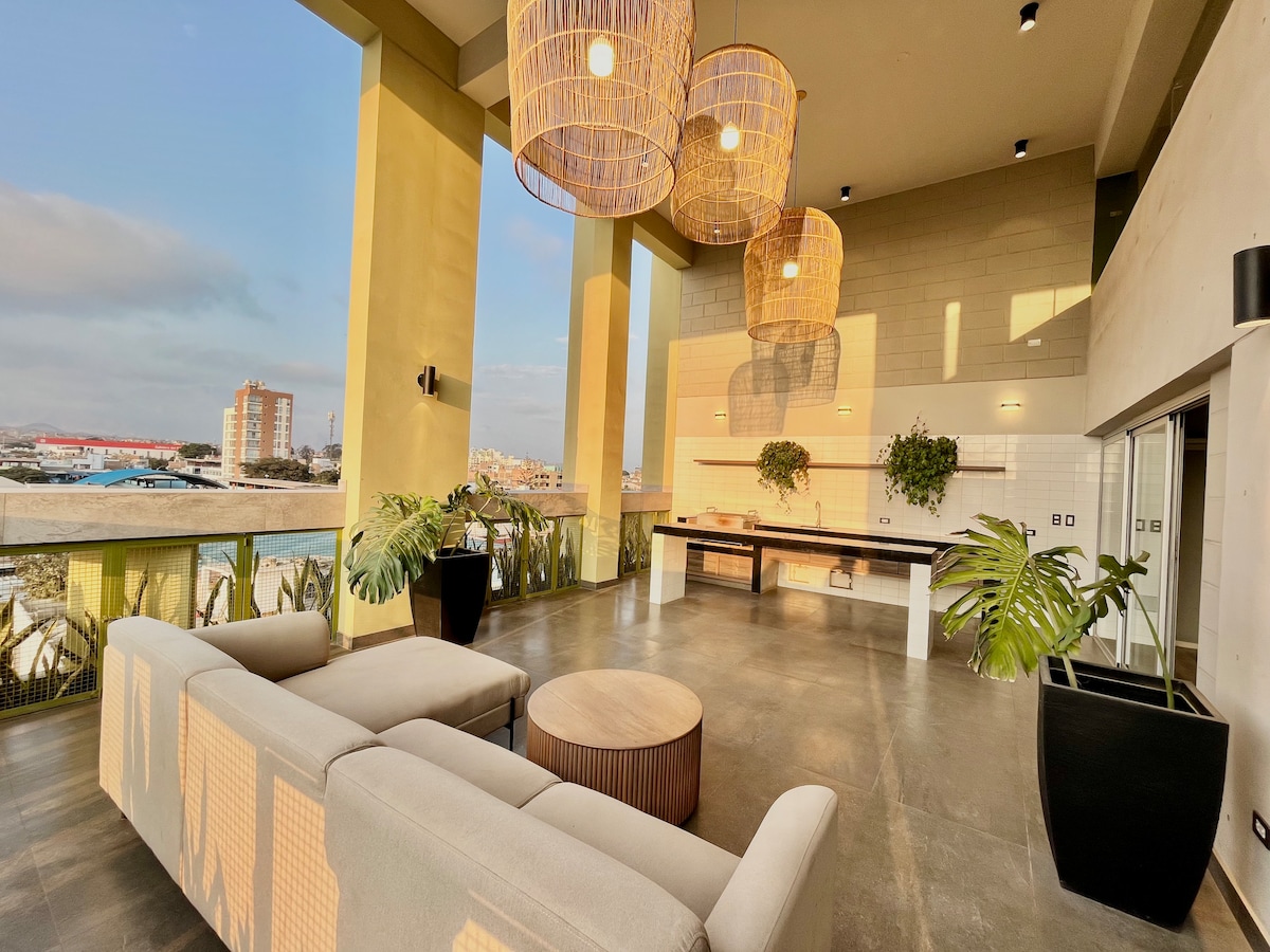 An open communal area is illuminated by oversized pendant lights, featuring a comfortable lounge setup with a light-colored sofa and round table. Large windows provide abundant natural light and views, while lush plants add a touch of greenery to the contemporary space.