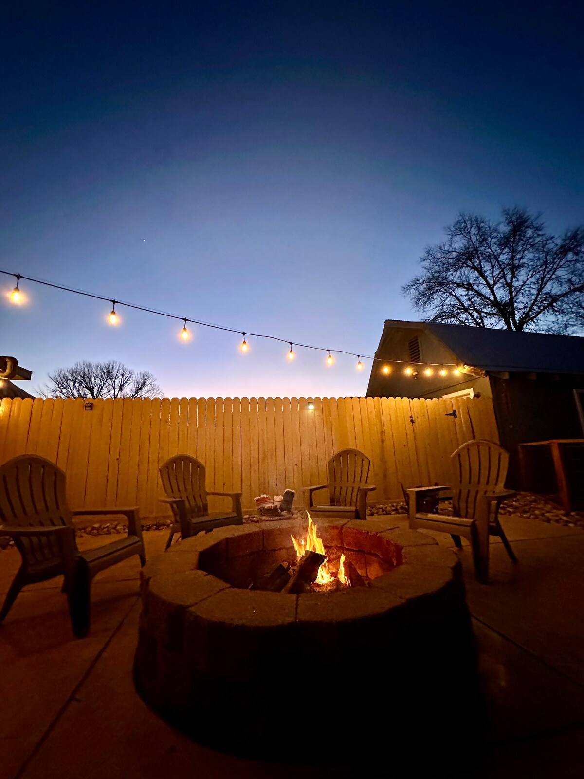 A cozy outdoor fire pit is surrounded by four wooden chairs, set against a backdrop of a wooden fence. String lights are gently illuminating the area, creating a warm ambiance as twilight descends in the sky.