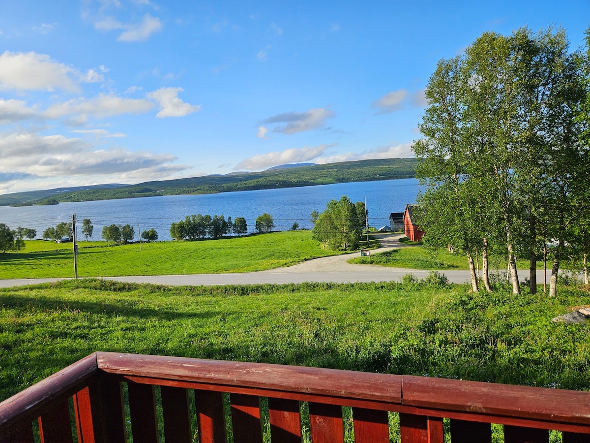 A view from the porch showcases the serene landscape, featuring a large lake with rolling hills in the background. Lush green fields and scattered trees complement the peaceful scenery, while a red building is visible in the distance.