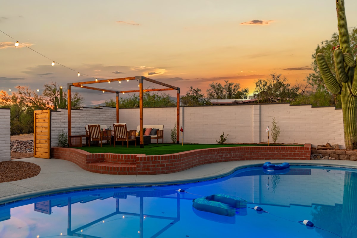 A serene outdoor space features a beautifully illuminated pool surrounded by desert vegetation and a scenic sunset view. A sturdy pergola with comfortable seating is visible, providing shade, while floaties gently rest on the water's surface.