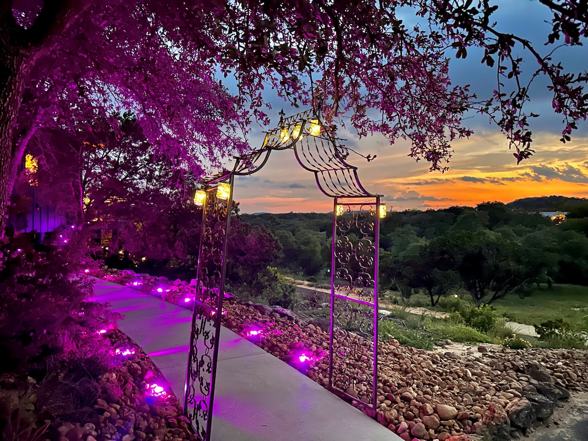 A decorative archway adorned with soft glowing lights frames a scenic pathway. The vibrant colors of sunset illuminate the landscape, emphasizing the natural beauty of the Texas Hill Country surrounded by lush greenery and a lightly textured stone pathway.