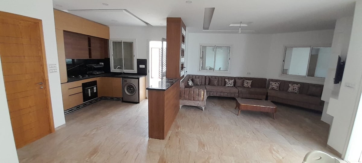 A spacious living area features a comfortable L-shaped sofa adorned with decorative cushions, complemented by a wooden coffee table. An open kitchen with modern cabinetry is visible, alongside large windows allowing natural light to fill the room. The flooring is finished with light-colored tiles.
