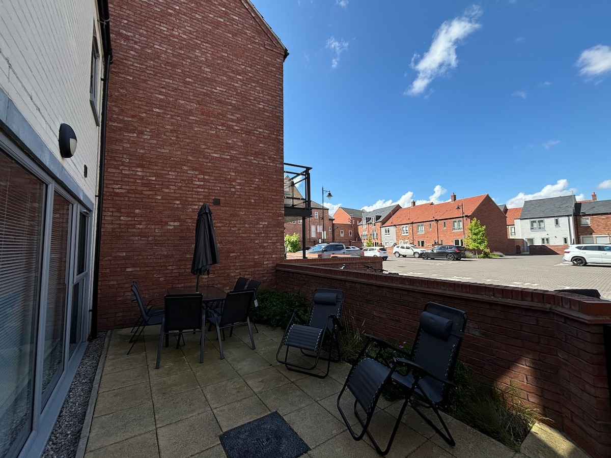 An outdoor terrace is presented with two loungers and a dining table set for four. A large umbrella provides shade. The surrounding area features a landscaped border, and a view of the parking area is visible, showcasing residential buildings under a clear blue sky.