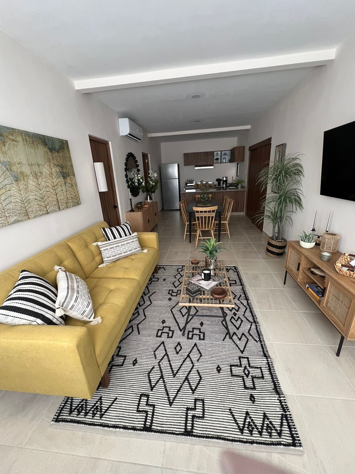 A modern living area features a yellow sofa adorned with patterned cushions, complemented by a geometric area rug. A dining table with wooden chairs is visible in the background, along with a kitchen area equipped with stainless steel appliances. Decorative plants add a touch of greenery.