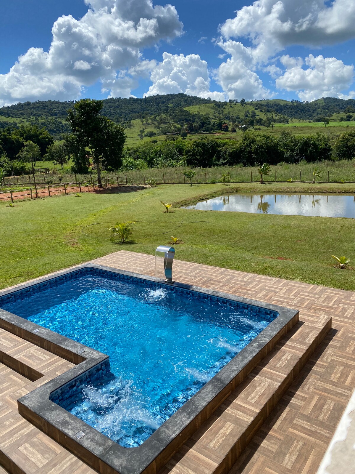 A private pool with clear blue water is visible, surrounded by a textured deck. Lush green hills and a clear sky with scattered clouds form the backdrop. An inviting open space with grass and a serene pond are seen nearby, enhancing the relaxing outdoor environment.