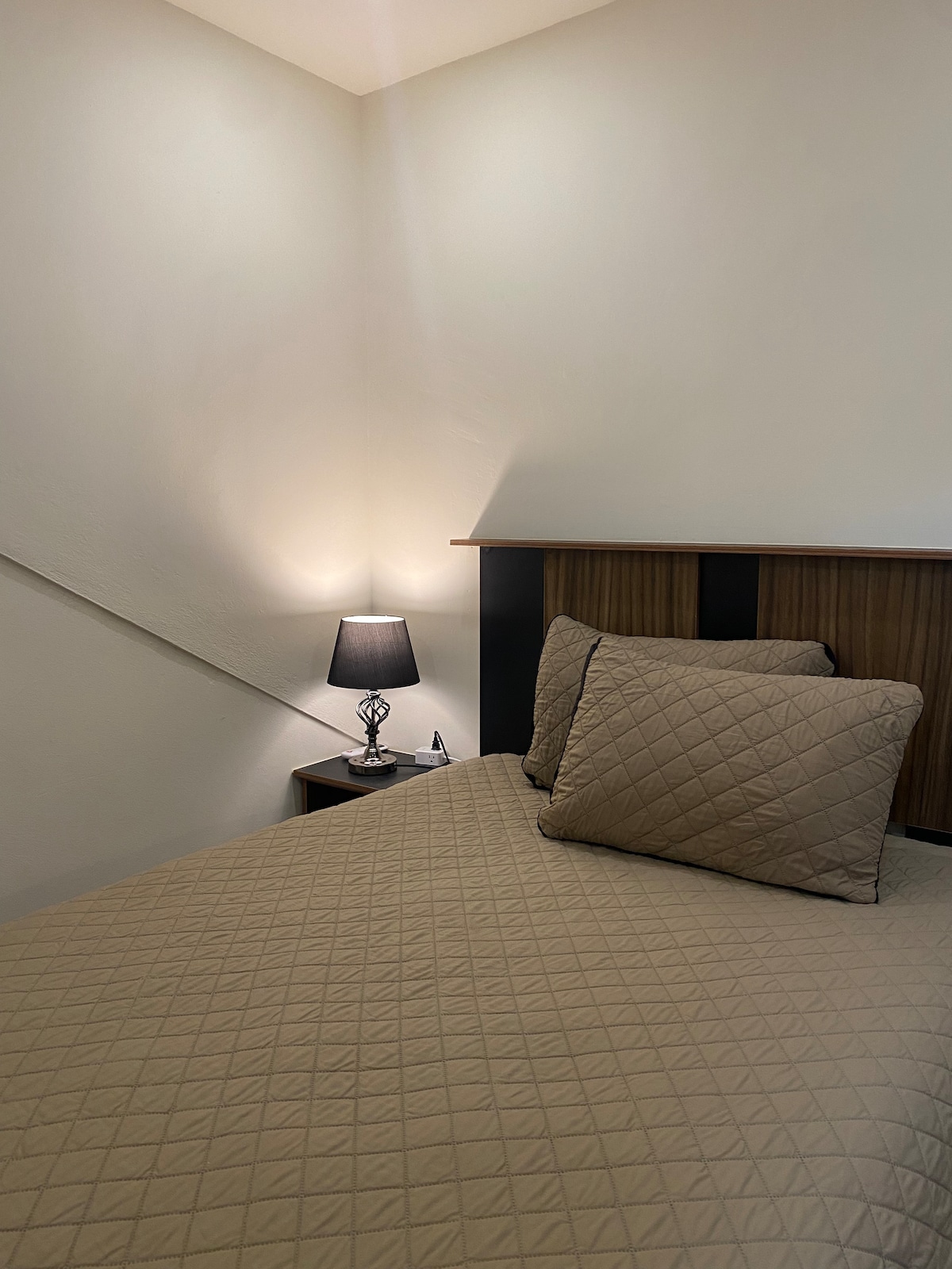 A cozy sleeping area features a spacious bed with beige quilted bedding and matching pillows. A bedside table holds a lamp, providing soft illumination. The room is accented by a minimalist wall design and a gentle slope visible in the background.