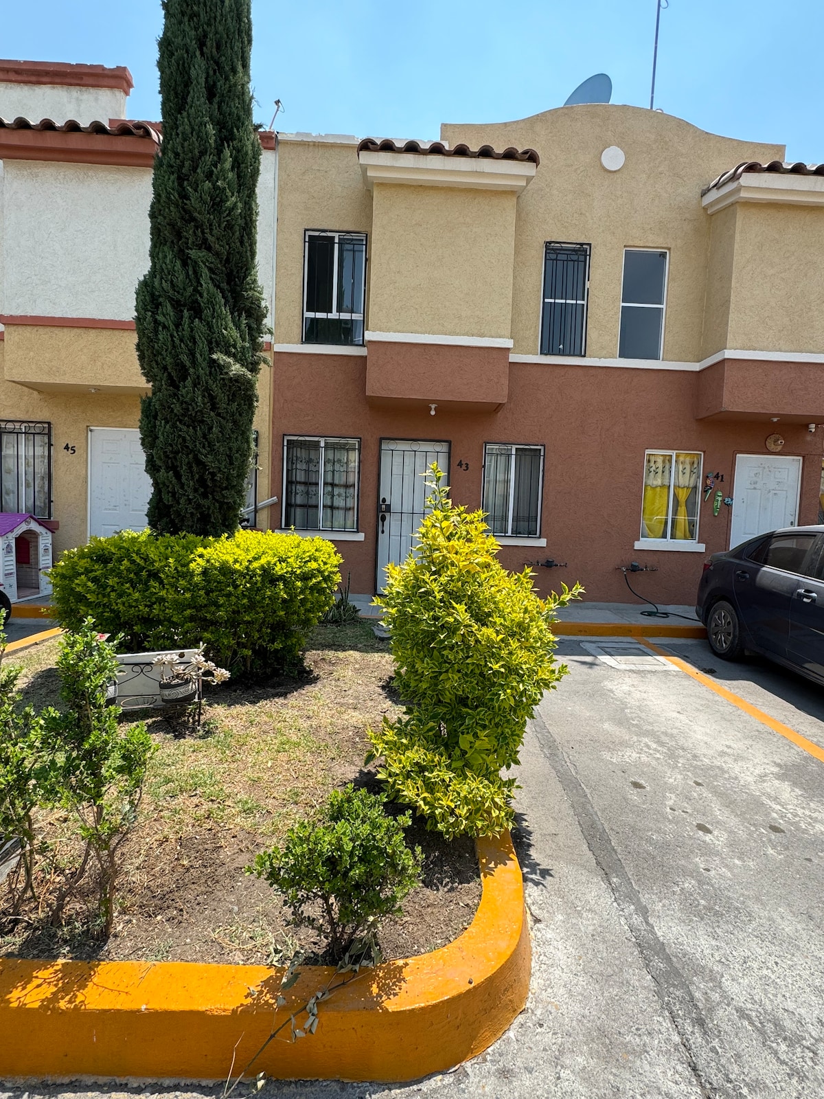 A two-story building with a combination of beige and terracotta-colored walls is depicted. Well-maintained shrubbery and a small garden area surround the entrance. Parking spaces are visible nearby, showcasing a clear pathway leading to the doors.