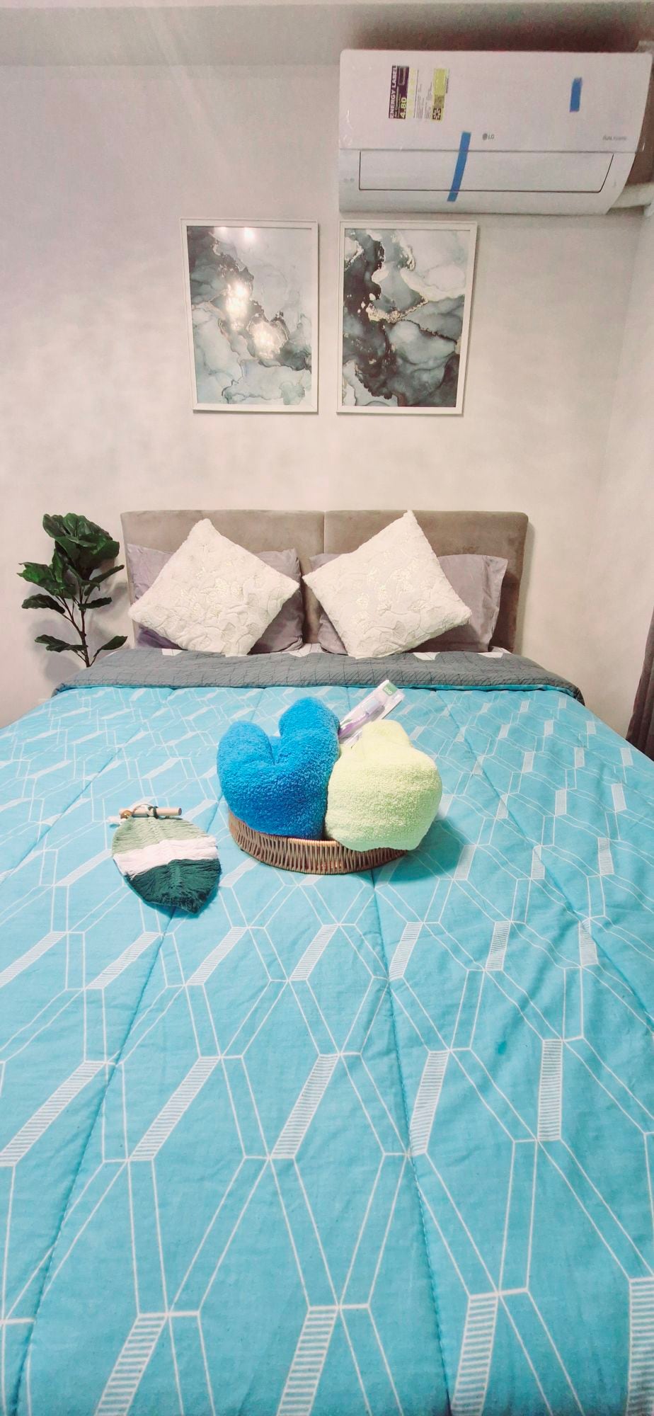 A comfortable bedroom features a spacious bed with a geometric-patterned turquoise blanket. Two decorative pillows in light gray sit atop the bed. Framed art pieces are displayed on the wall above the bed, complemented by a potted plant in the corner.