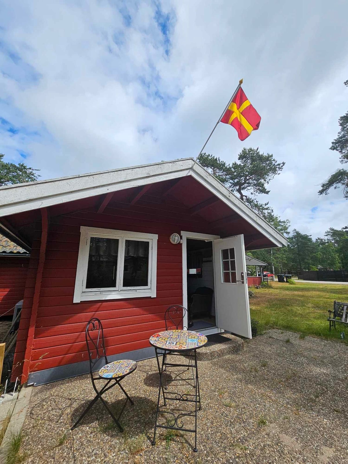 A red wooden cottage is depicted with a bright flag flying above. A small outdoor table with decorative tiles and two iron chairs is visible in front of the entrance. The surrounding grassy area and trees provide a natural setting.