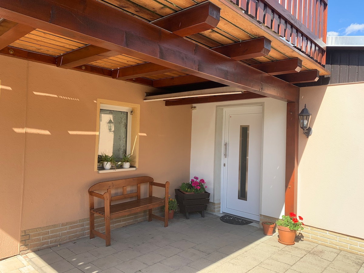 An entrance area features a modern front door framed by a welcoming overhang. A wooden bench provides seating, while potted plants add a touch of greenery. The space is illuminated by natural light, contributing to a cheerful and tidy appearance.