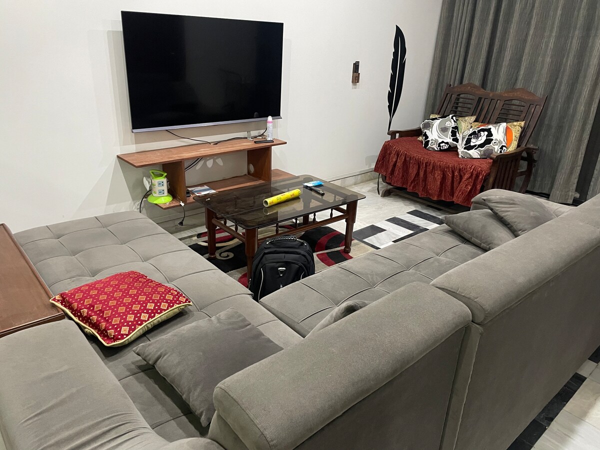 A comfortable living area is showcased with a sectional gray sofa and a wooden coffee table. A television is mounted on the wall, and a vibrant throw pillow adds a pop of color on the sofa. Wooden seating is also visible, combined with decorative curtains.