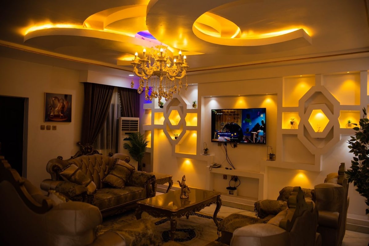 A stylish living room is highlighted by elegant lighting and ornate decor. A comfortable seating area is arranged around a central table, while a wall-mounted television serves as a focal point. A chandelier adds a touch of sophistication, complemented by contemporary architectural features.