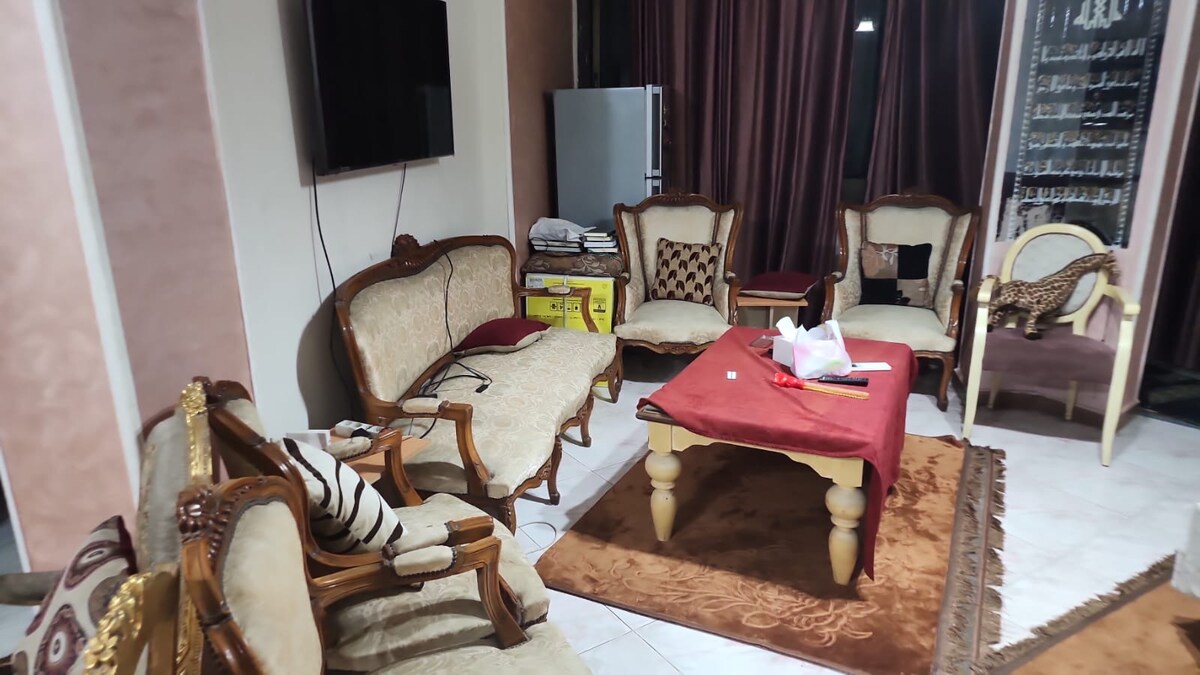A comfortable living area contains a set of upholstered seating, including chairs and sofas arranged around a low table. A TV is mounted on the wall, and a large window is draped with thick curtains. A decorative rug covers the tiled floor.