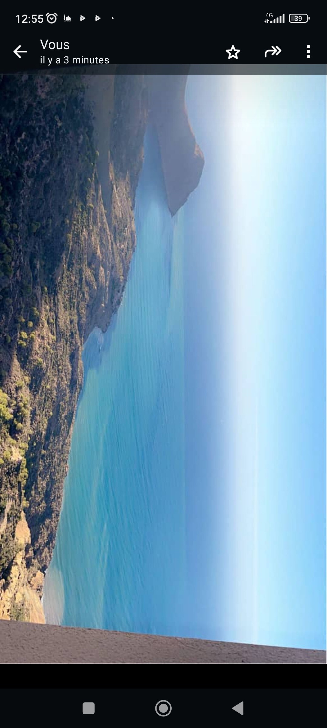 A panoramic view of the coastline reveals calm turquoise waters meeting rocky formations and gentle hills in the distance. The sky is a clear gradient from blue to light, creating a serene backdrop for the natural landscape.