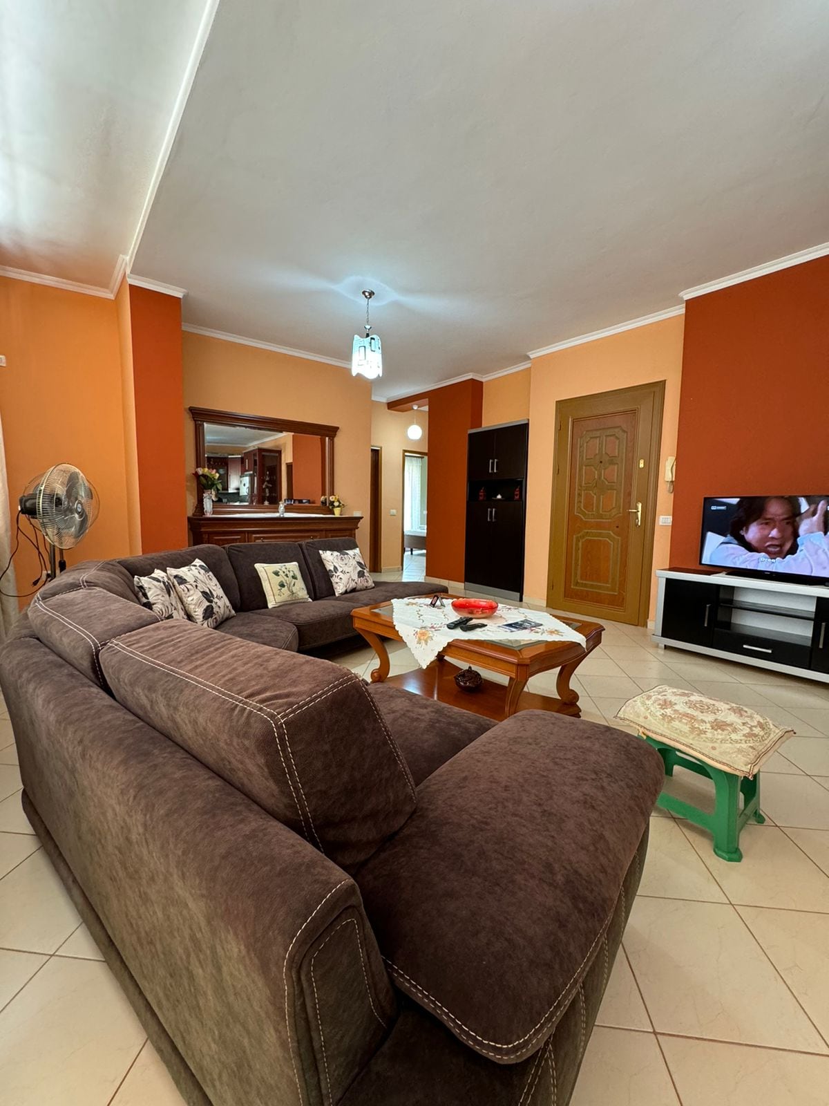 A spacious living area features a large, plush sectional sofa facing a modern television. Natural light is drawn in through nearby windows, and warm orange walls enhance the cozy ambiance. A wooden coffee table and decorative accents add to the inviting decor.