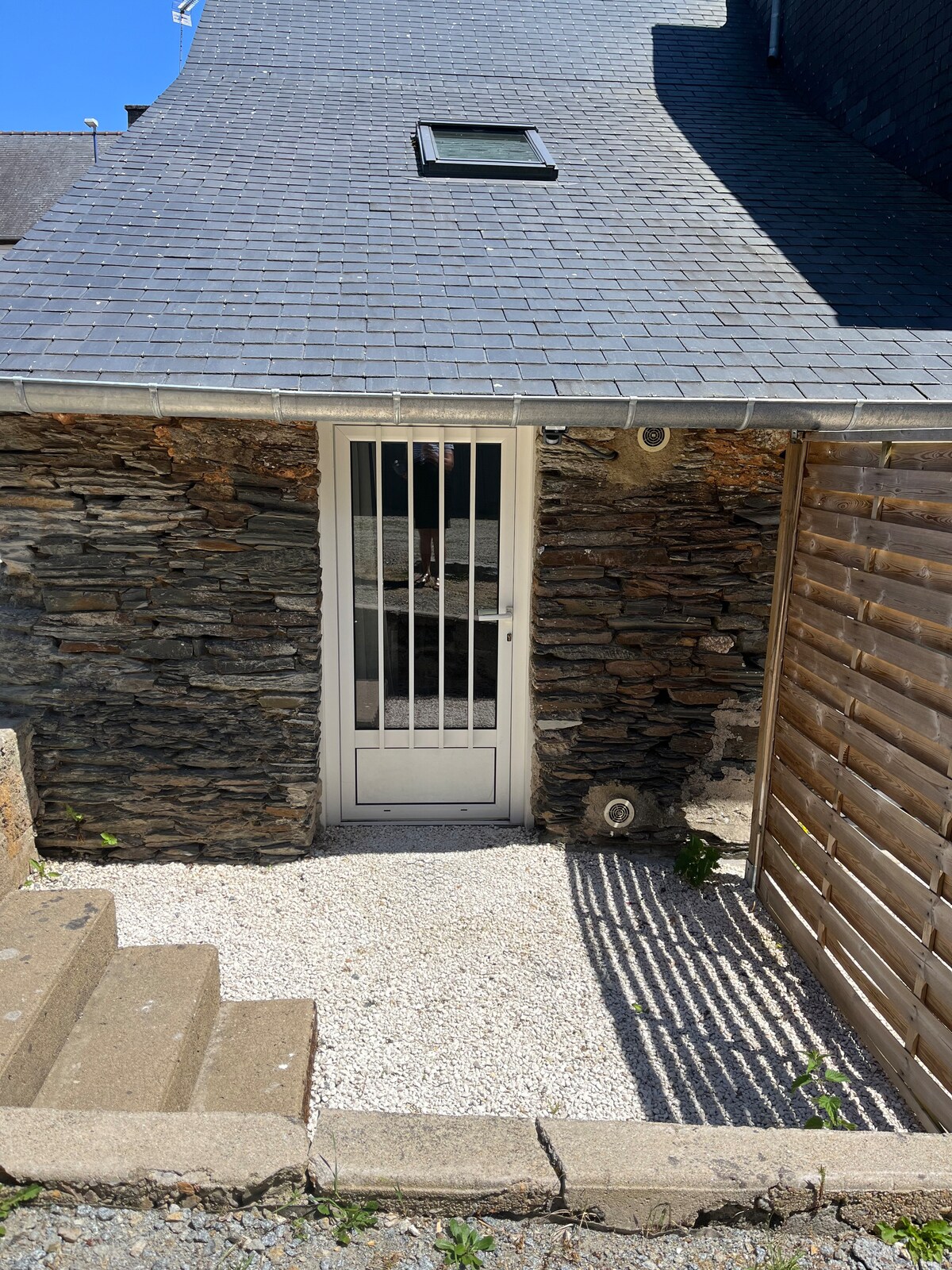 An entrance is framed by rustic stone walls and features a white door with security bars. Steps lead up to the entrance, which is surrounded by small gravel stones. Sunlight casts a shadow onto the pathway, enhancing the welcoming appearance.