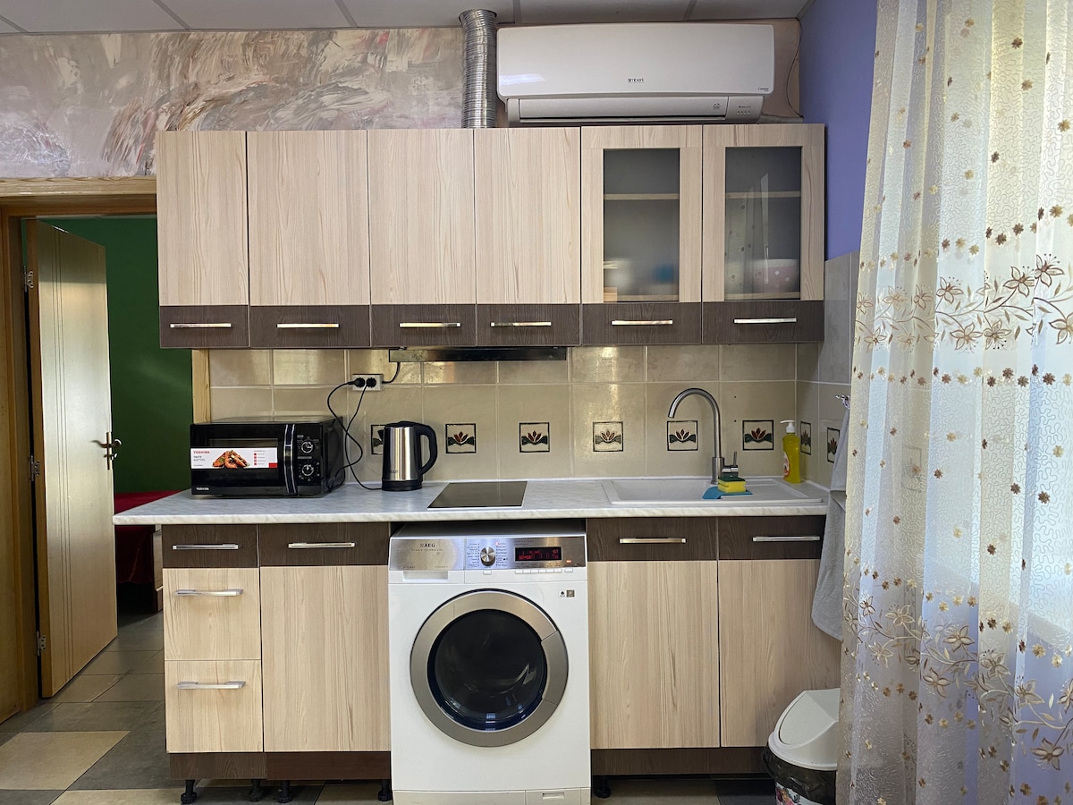 A functional kitchen area features wooden cabinetry with a combination of light and dark tones. Appliances including a microwave, kettle, and washing machine are visible. A window draped with a sheer curtain allows natural light to enter, enhancing the space's brightness.