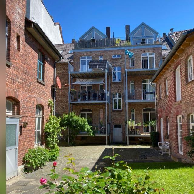 A tranquil courtyard showcases a three-story brick building, featuring multiple balconies adorned with plants. Light reflects off the red brick facades, and the garden area is lined with greenery, creating a serene outdoor space. The clear blue sky enhances the inviting atmosphere.