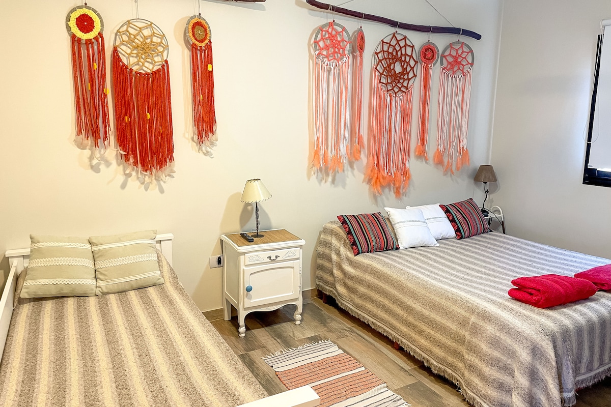 A cozy bedroom is presented with two queen-size beds, each adorned with soft linens and vibrant blankets. Colorful dreamcatchers hang on the wall, adding a decorative touch. A bedside lamp and a small table offer functional comfort, while natural light filters through the window.