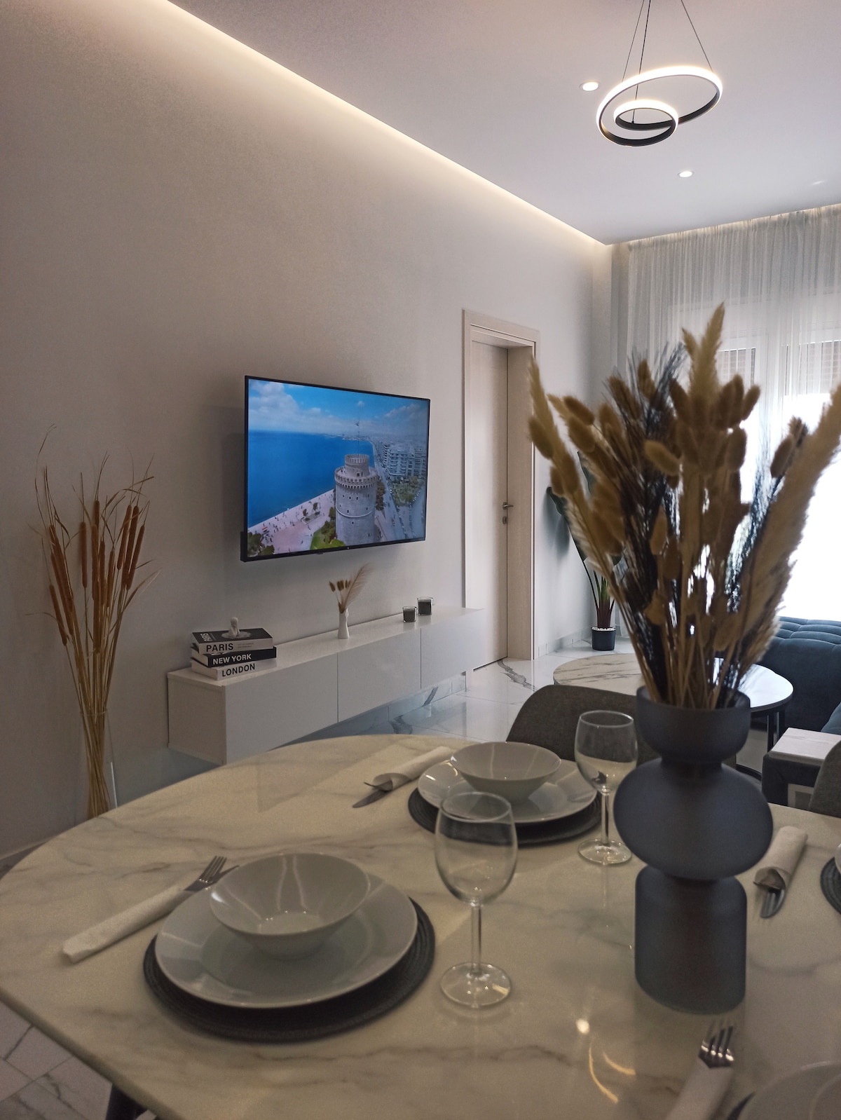 A modern dining area is displayed, featuring a marble table set with elegant dishware and crystal glasses. A large screen TV mounted on the wall shows a scenic view, while decorative vases with dried grass add a touch of sophistication to the space.