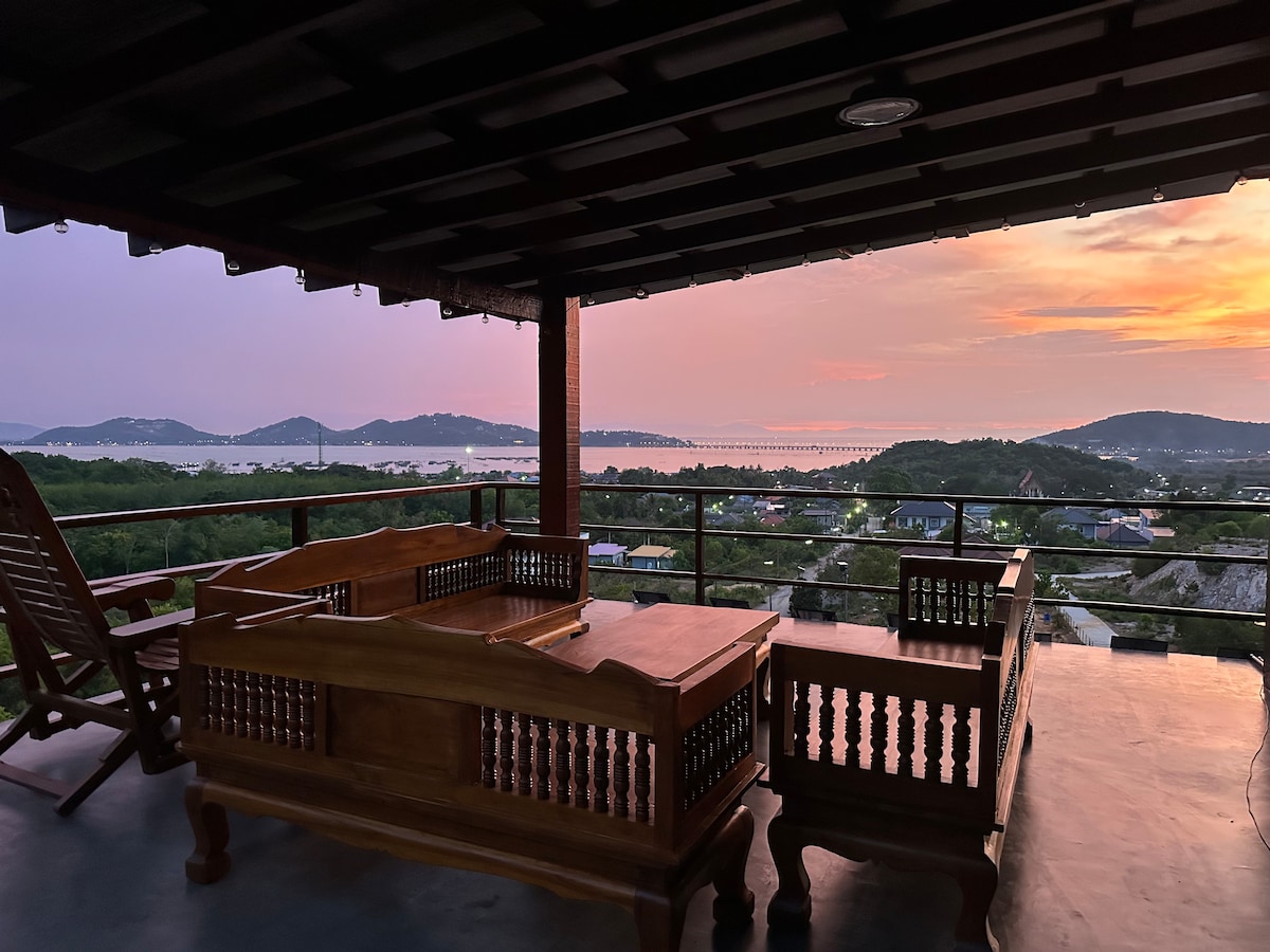 A spacious outdoor deck features a wooden seating area with decorative detailing, offering views of the sea and hills. The sunset casts soft hues across the sky, creating a serene backdrop for relaxation. The area is partially shaded, enhancing comfort.