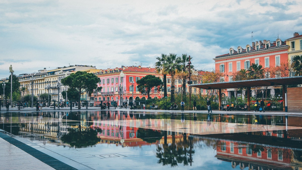 A vibrant scene reflects off calm water, capturing colorful buildings along the promenade. Palm trees frame the view, contributing to the lively atmosphere of the area. People can be seen strolling, enjoying the outdoor space and the serene setting.